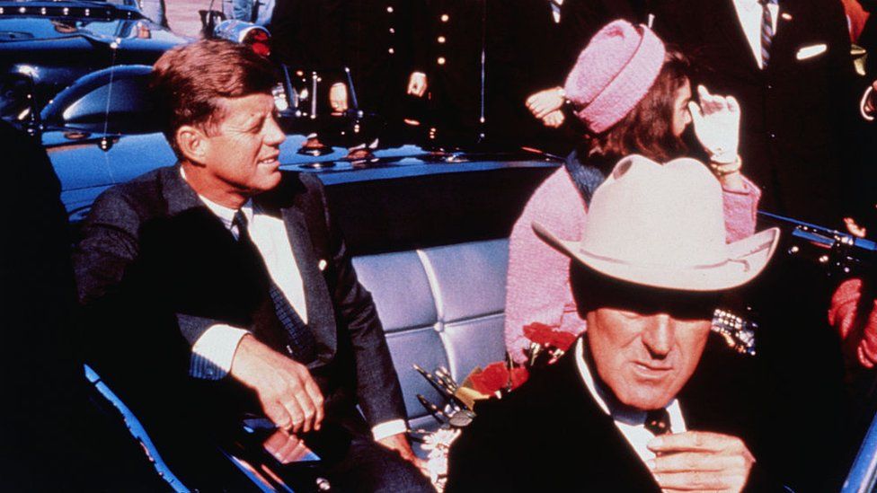 Texas Governor John Connally adjusts his tie (foreground) as US President John F Kennedy (left) & First Lady Jacqueline Kennedy (in pink) settled in rear seats, prepared for motorcade into city from airport, Nov. 22. After a few speaking stops, the President was assassinated in the same car.