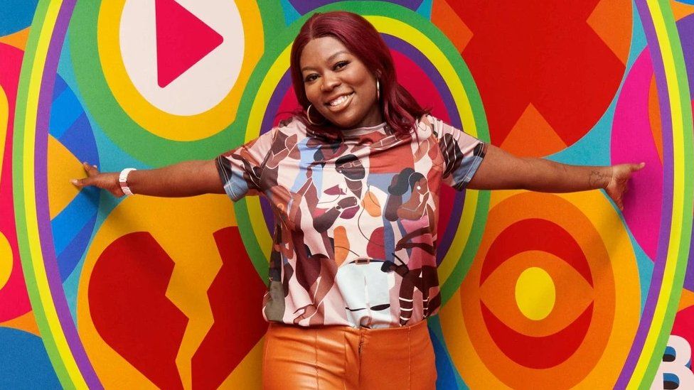 Big Brother contestant Trish wearing orange trousers and a colourful top with people on it, in front of a multicoloured background