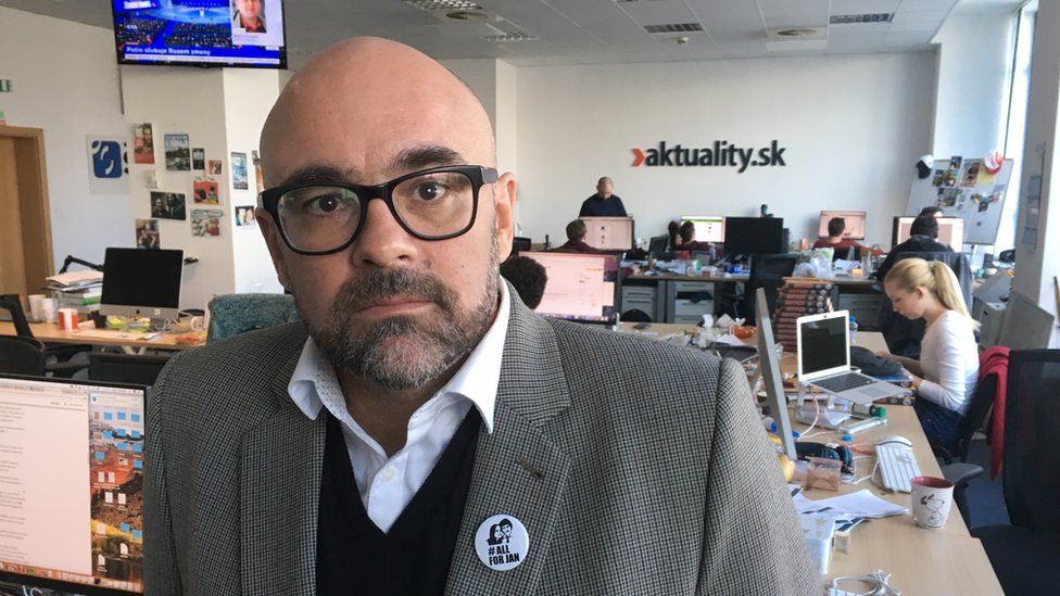 Peter Bardy, a man with a close-cropped beard, wearing a business casual shirt and jacket combination, looks into the camera with the offices of aktuality spreading out behind him, with journalists at work at their desks