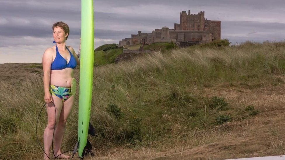 Gill stands with a surfboard with Bamburgh Castle beyond