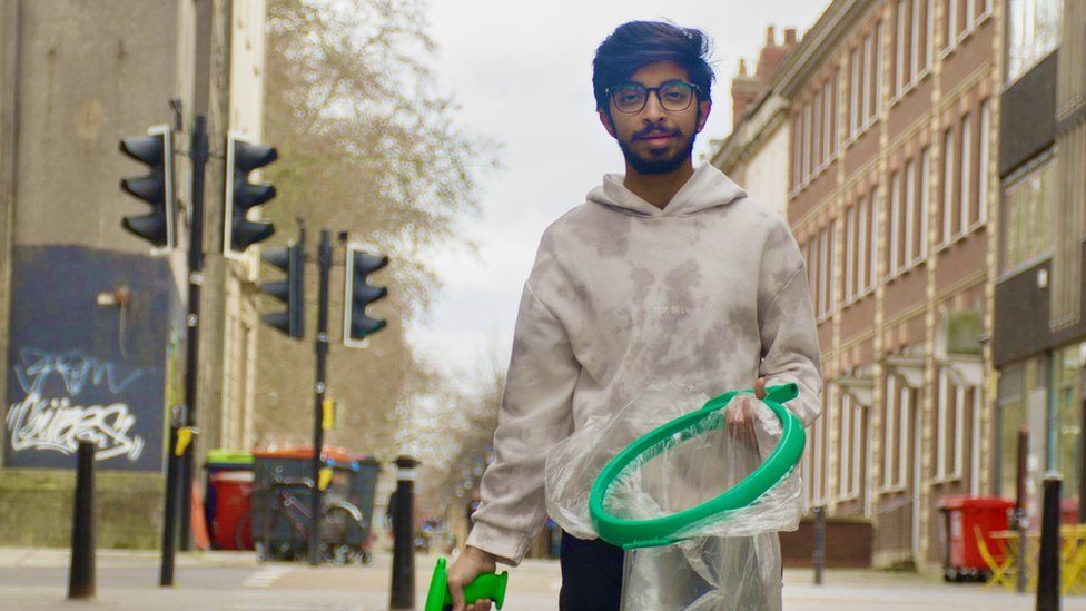 Vivek is stood up holding a litter picker and bag in a built-up area of Bristol