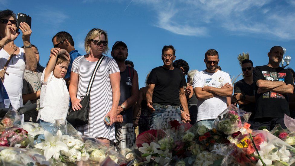 People stand near a makeshift memorial for the victims killed in an attack where a truck mowed through revelers in Nice, southern France, Friday, 15 July 2016.