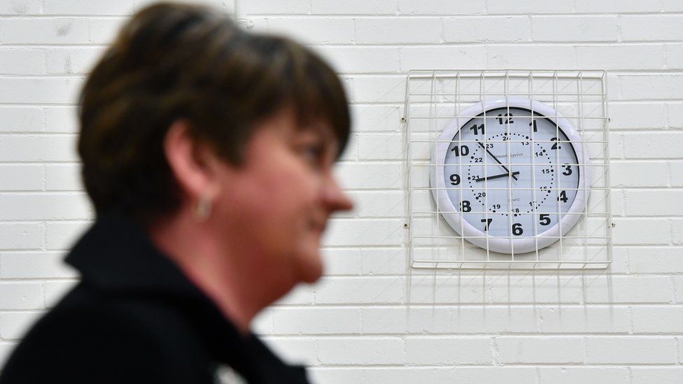 Arlene Foster stands in front of a clock on a wall