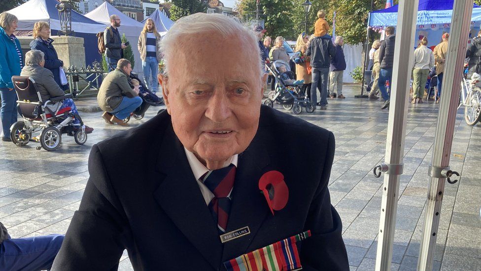 Ron Collins sitting down in a suit wearing a poppy and his medals, with Salisbury market square in the background full of people