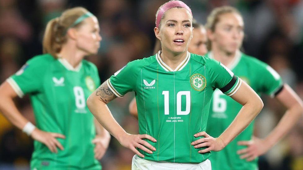 Republic of Ireland player Denise O'Sullivan stood on the pitch during their Women's World Cup game against Australia. She is stood with her hands on her hips and is wearing a green Republic of Ireland football kit with the number 10 on the front. She also has a pink headband in her hair. Behind her and out of focus there are 3 other Republic of Ireland players also wearing the green kit and with their hands also on their hips.