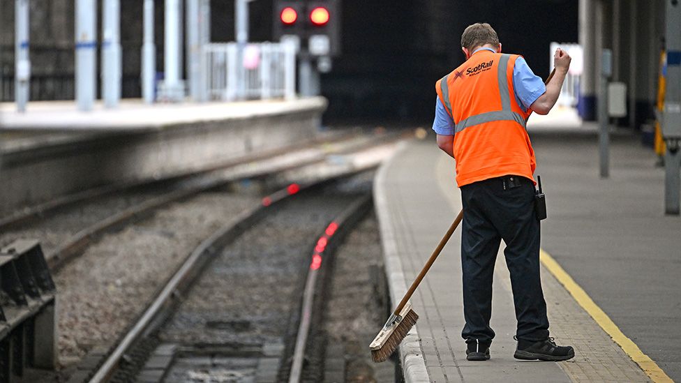 A rail worker sweeps the platform at a train station next to the train tracks