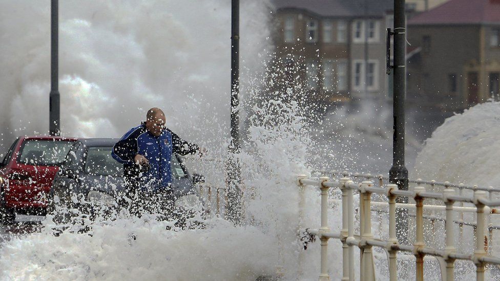 A man gets caught in spray as hide tide breaches the sea wall throwing up debris and flooding the main road in the coastal village of Carnlough, in Northern Ireland