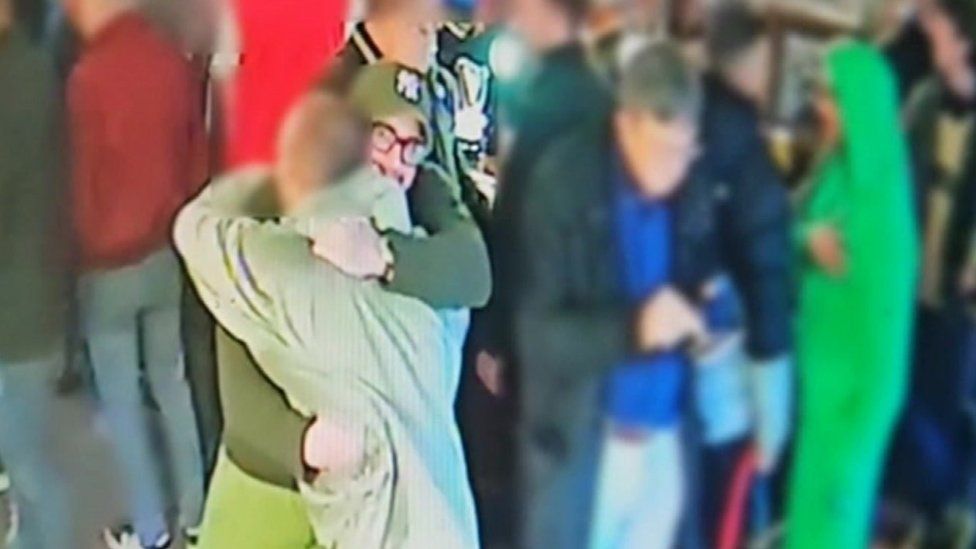 CCTV image of a man with a hat and glasses hugging another man, both dressed in green