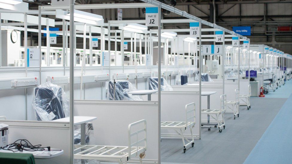 Rows and rows of hospital beds are seen, with parts still wrapped in plastic, in a cavernous space