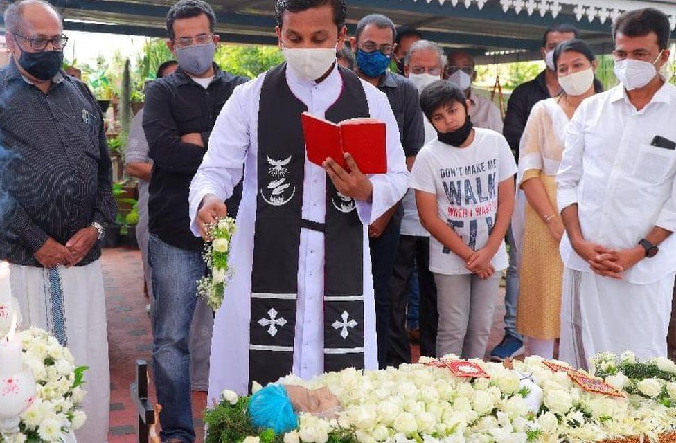 A priest sprinkles holy water on the deceased as relatives watch one