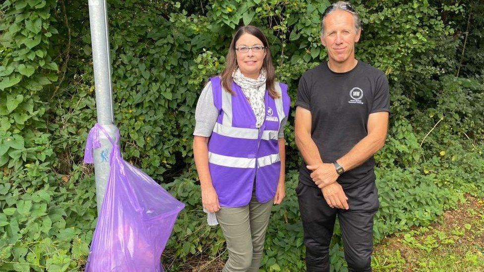 Woman in hi-viz jacket and man in black T-shirt stand next to rubbish bag