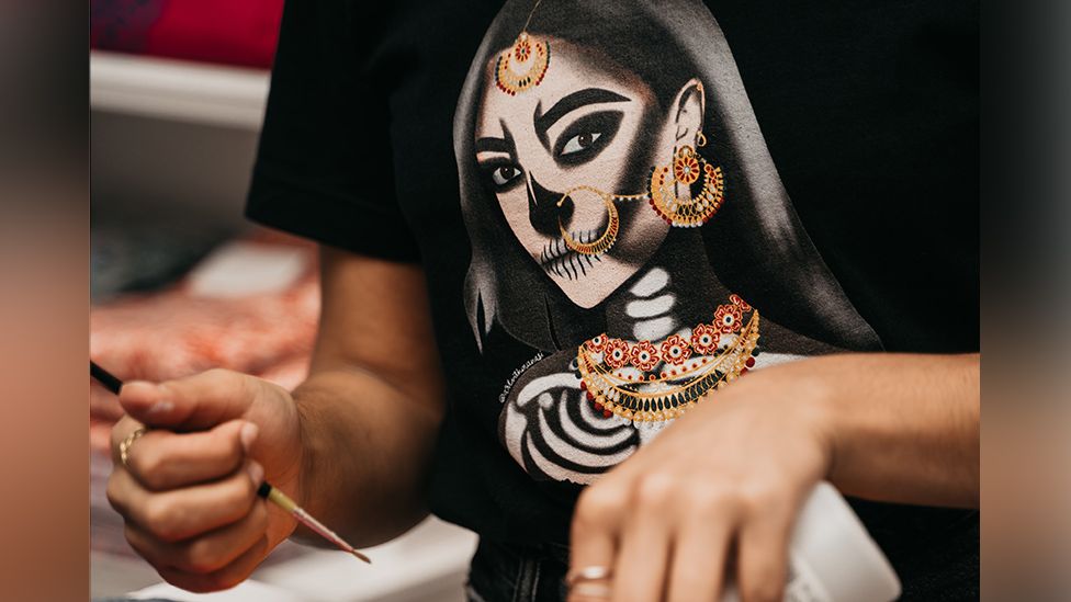 A person doing some sort of arts and crafts holds a paintbrush in one hand and a squeezy bottle of paint or glue in the other. She's wearing a black t-shirt with a design showing a South Asian woman wearing traditional dress - a headscarf can been seen and she's wearing elaborate jewellery on her forehead, ears and neck. However, the female figure is a skeleton rather than human - we can see her ribs, and her features are heavily stylised.