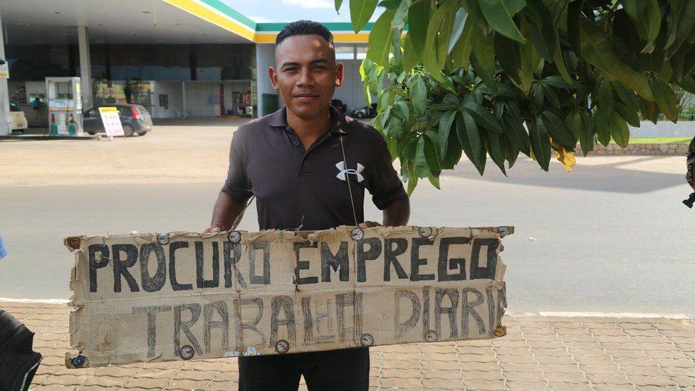 A man carries a sign reading "Looking for work" in Portuguese around his neck in Boa Vista