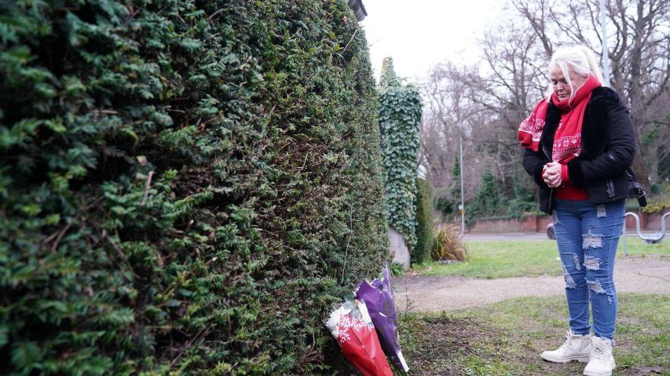 Member of the public in a black jacket and wearing a red festive scarf leaves flowers outside Wensum Park
