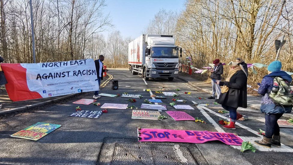 Flowers and placards laid on road with campaigners stood around outside centre