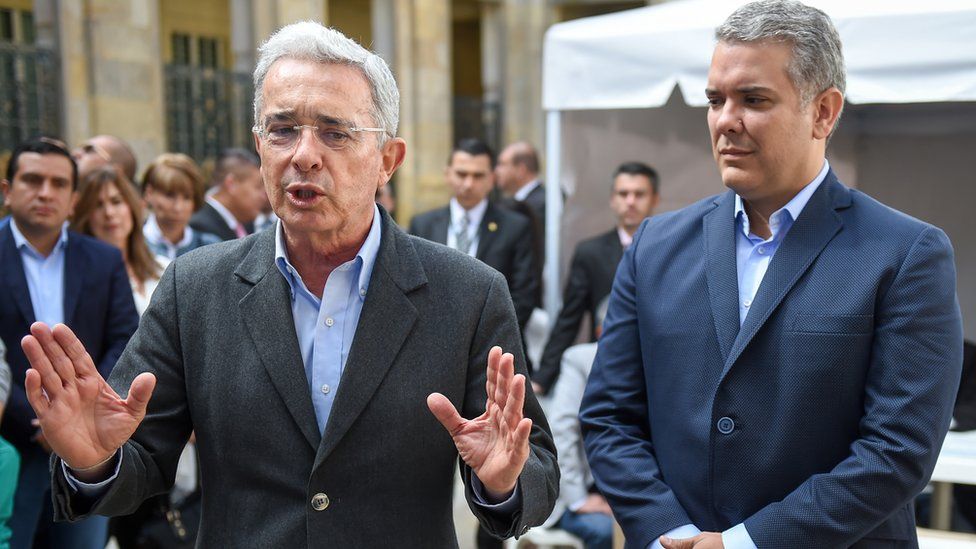 Mr Uribe gestures with open palms next to a younger Mr Duque, standing to attention nearby