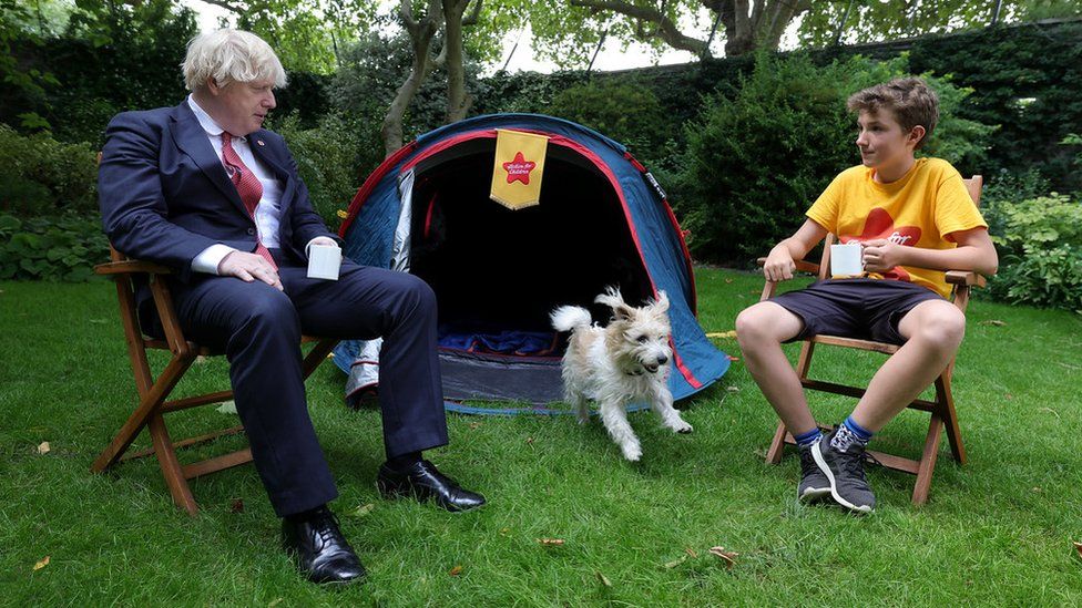 Boris Johnson and Max Woosey sit next to tent as Dilyn the dog runs past them