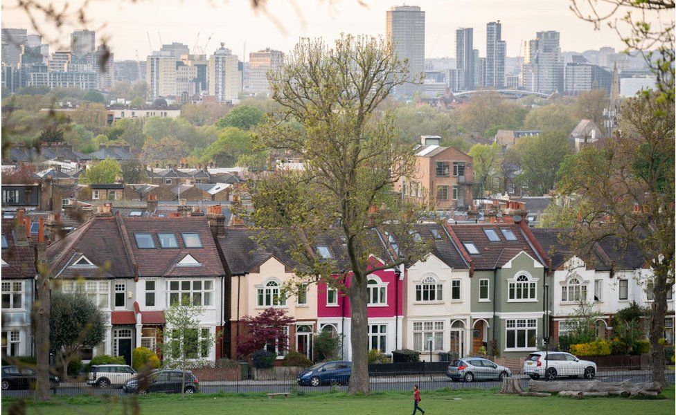 Through a gap of 100 year-old ash trees are Edwardian period homes bordering Ruskin Park in south London with residential homes and businesses in the distance, on 19th April 202,