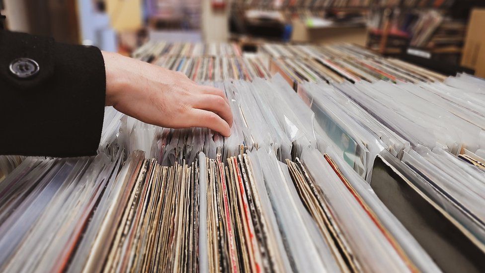 A close-up view of a tightly packed rack of vinyl records. Many are covered in a plastic protective sleeve, but some have the dog-eared ends of their cardboard covers exposed. A hand is seen flicking through the different titles. In the background the wider shop can just be made out, with shelves and boxes packed with records visible. The shop has a thrown-together look.