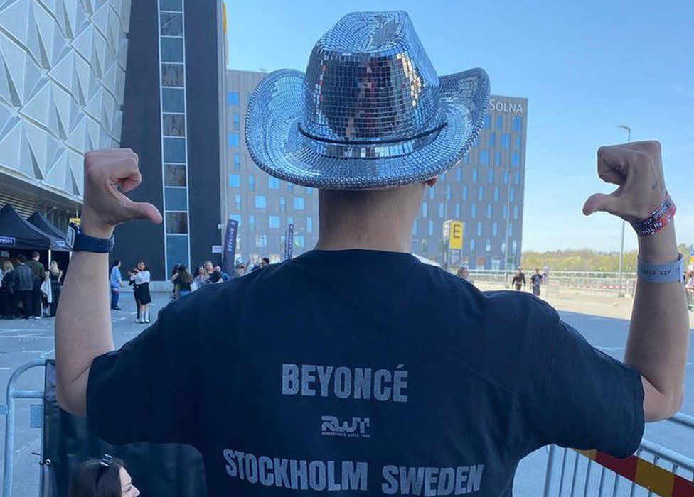 Bilal Rouabah with his back to the camera. He has both his hands up with his thumbs pointing down to the words on his black T-shirt which says andquot;Beyoncé" and then says "Stockholm Sweden" underneath it. He is also wearing a shiny silver cowboy hat.