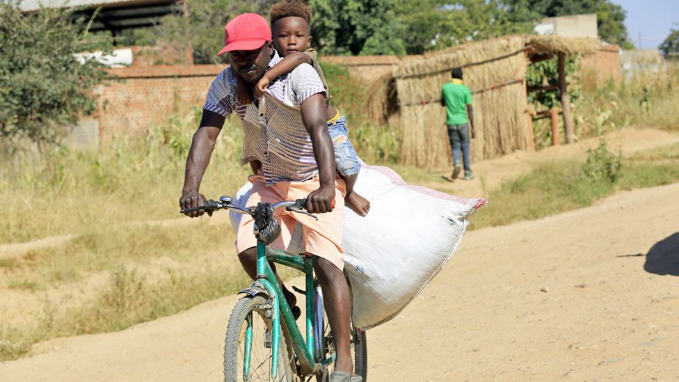 A man with a child o his back cycling in Epworth, Zimbabwe - Tuesday 31 March 202