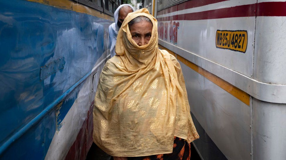 An old woman without wearing a safety mask covered her face with a cloth, crossing through the lane of a city bus terminus in Kolkata, India on July 1, 2021. West Bengal Govt has given certain restrictions on the lockdown effective from 1st july to 15th july. Bus, auto and toto services has been resumed with 50% capacity. Saloon, parlour and gym can be opened with 50% customers. Local train and metro will not be operational as per the Indian media report. Though the bus services were not regular as bus service unions protest to hike the bus fare and appealed for compulsory vaccination for all bus service operators. (Photo by Dipayan Bose/NurPhoto via Getty Images)