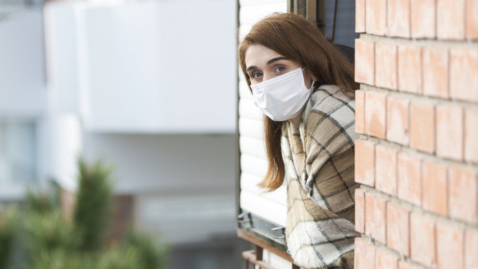 Woman wearing a mask looks out a window at the camera