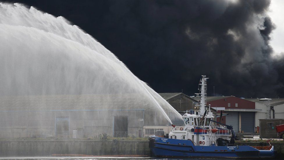 A firefighting boat sprays large streams of water towards a factory building that gives off thick black smoke.