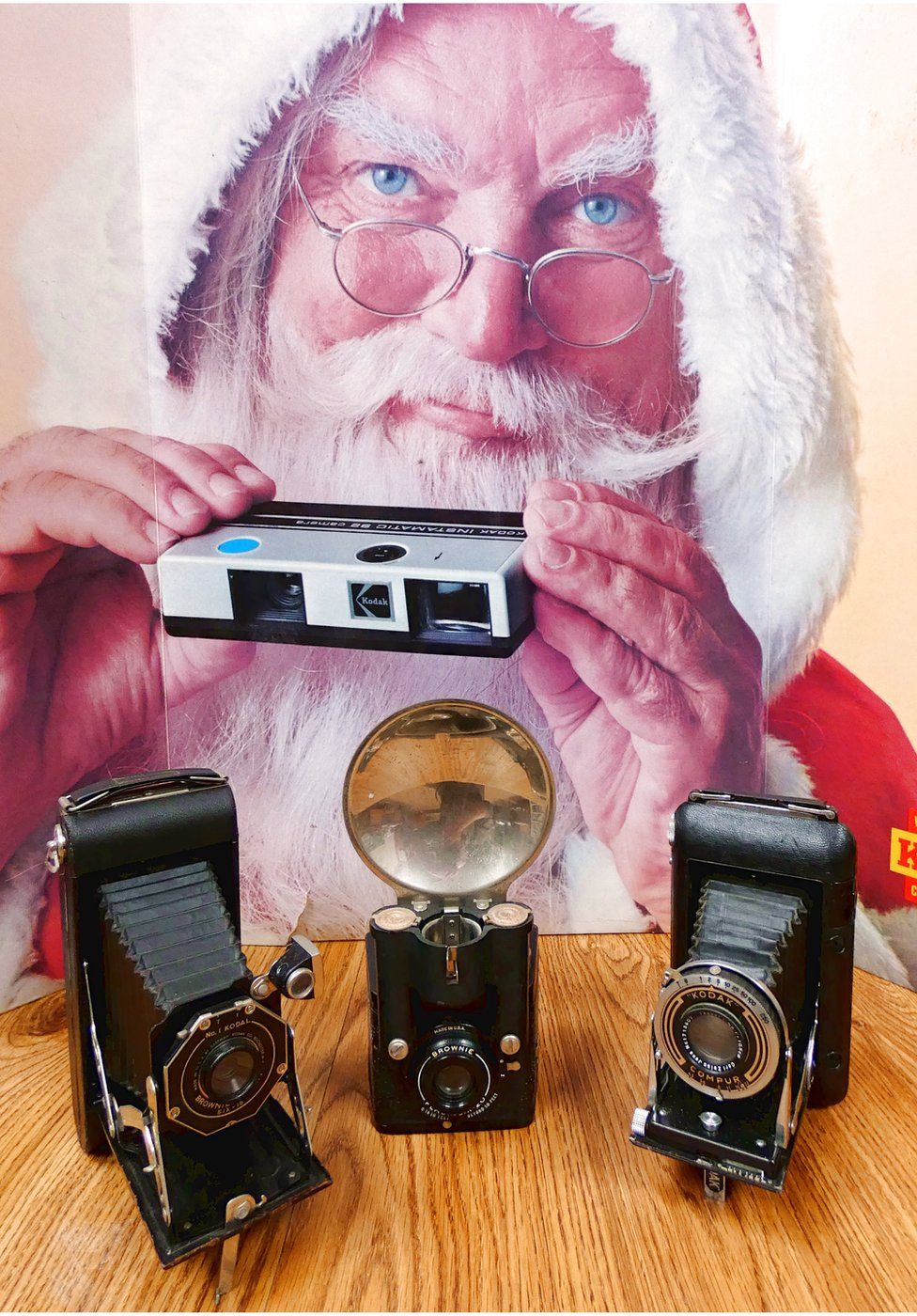 backdrop: advert for Kodak instamatic camera. Foreground, left to right: No.1 Kodal, Brownie Pliant, Six-16 folding camera (USA); Brownie Flash, Six-20, c.1941; Kodak Regent, Compur shutter, Zeiss Tessar lens, folding camera, c.1935-39 (Germany)