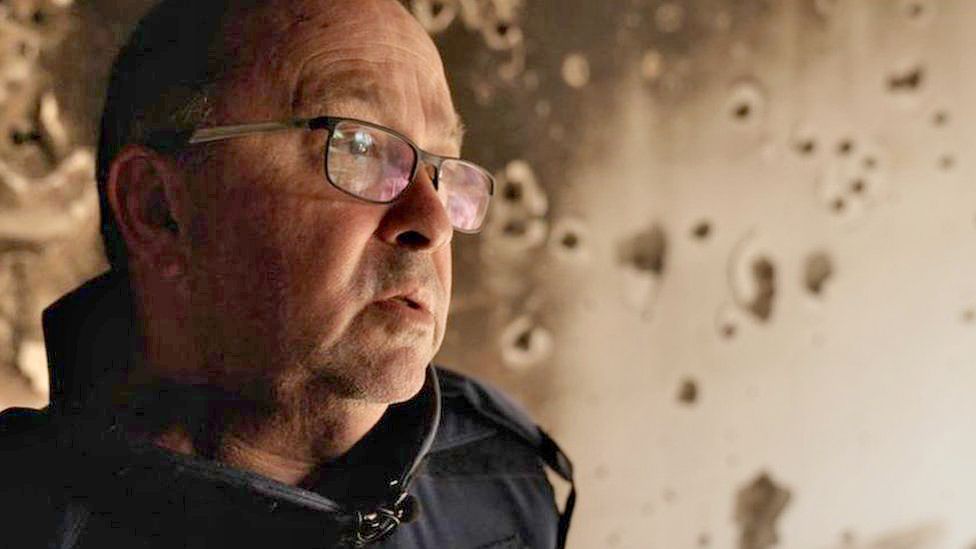 A man in glasses stands in front of a scorched wall marked with bullet holes.