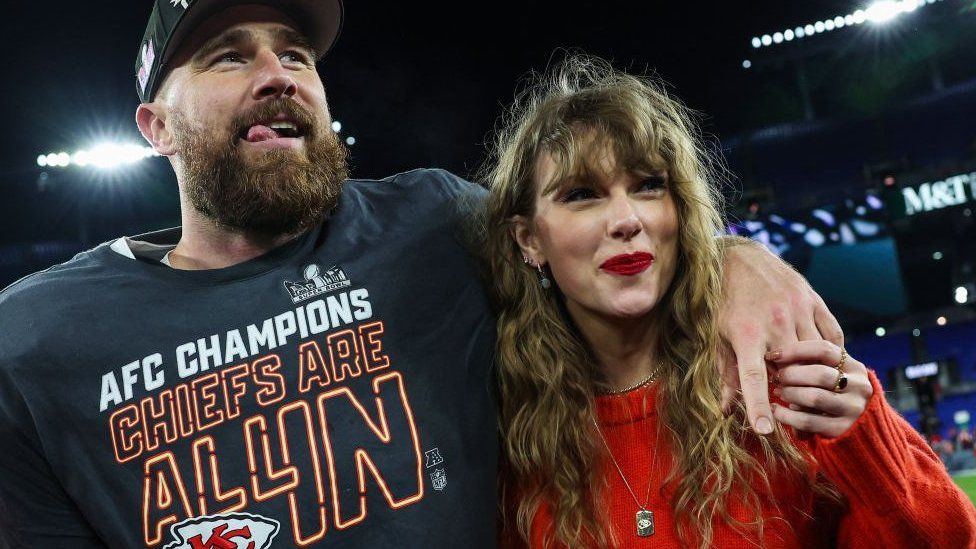 Travis Kelce and Taylor Swift, he is wearing American Football uniform and a hat and has his arm around her - she is wearing a red top and smiling