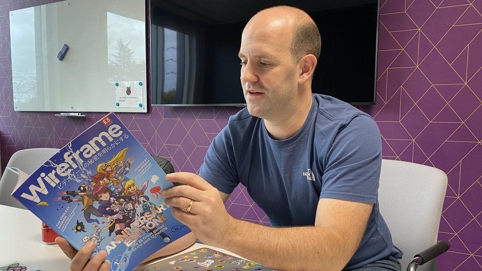 Eben Upton in an office wearing a blue t-shirt and reading a computer magazine