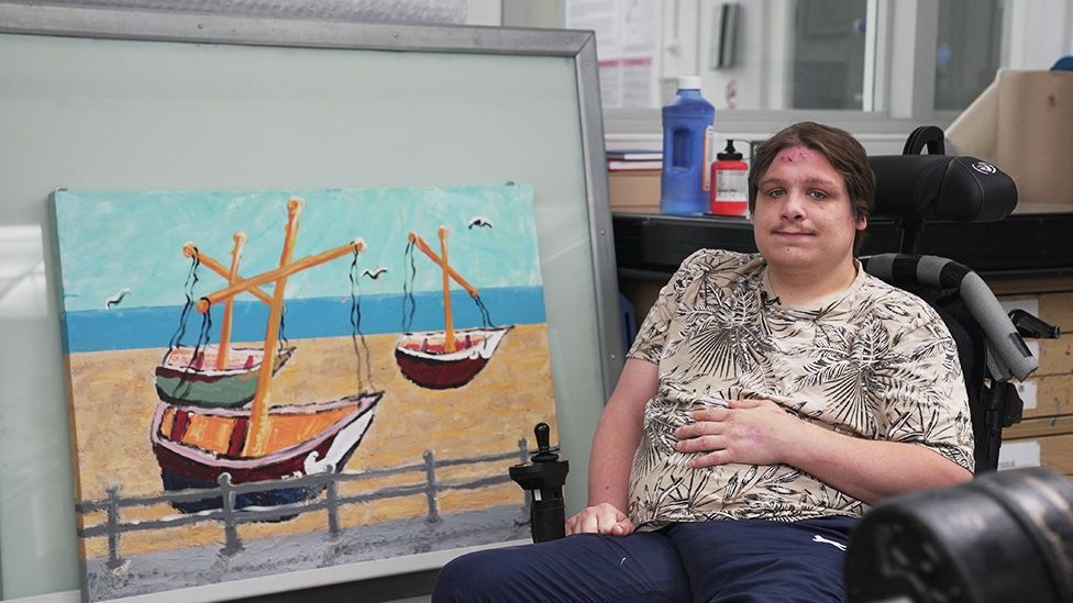 Matthew sitting alongside one of his paintings - a picture of boats on the beach