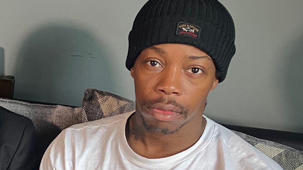 Head and shoulders image of Corey Wilson wearing a black beanie hat and white t-shirt sitting on a sofa