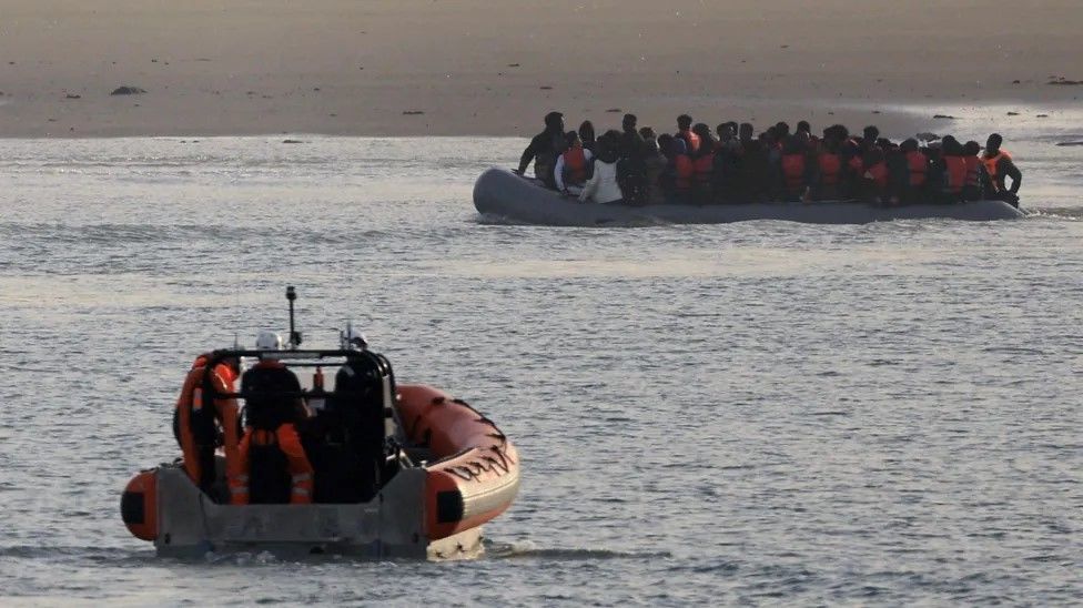 An orange inflatable French rescue vessel heading towards a large group of migrants on an inflatable dingy just off a sandy beach