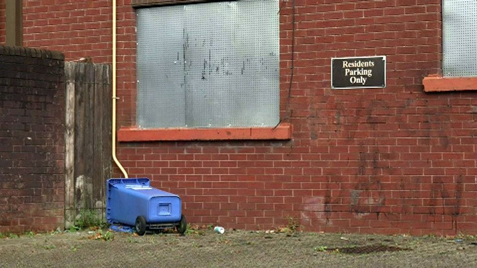 A boarded up red-brick terraced house. Two windows on the ground floor are boarded up. Blue wheelie bin on the floor. Parking sign in between boarded up windows. Aerial and top windows in view of shot.