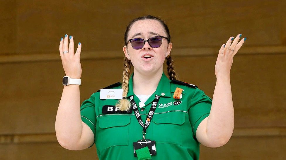 Hafwen looking at the camera. She has sunglasses on and a green St John Ambulance shirt on along with a lanyard. He hands are in the air as she is signing