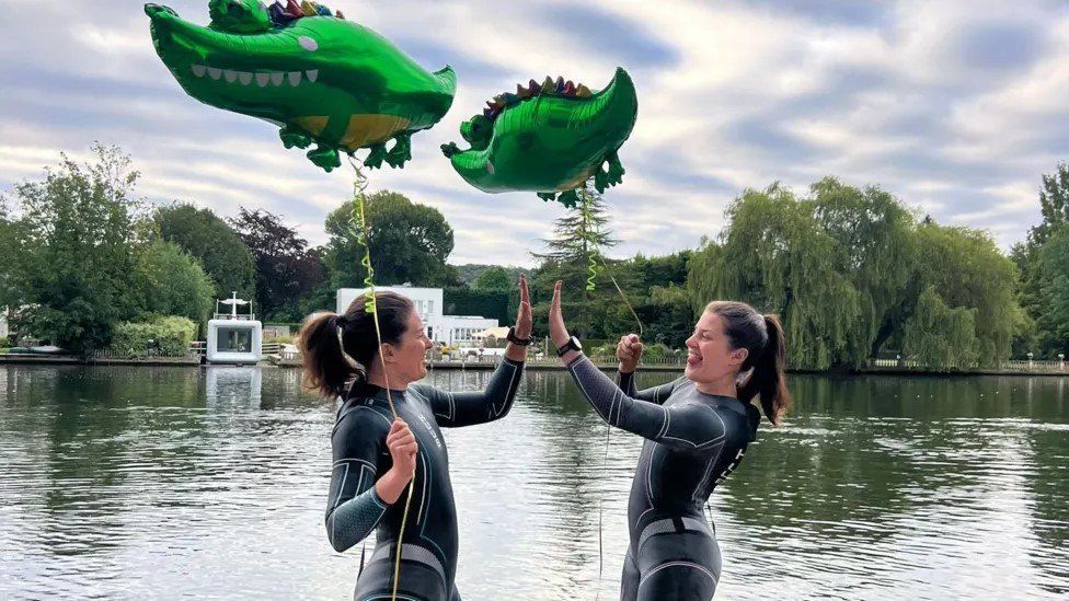 The twins high five each other in wetsuits holding crocodile-shaped balloons with the River Thames behind