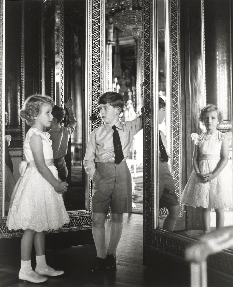 Princess Anne and King Charles as young children, standing beside a mirror in Buckingham Palace