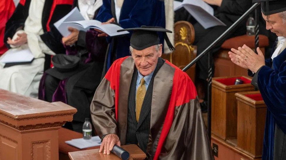 Sir Michael Palin in his university robes, holding his degree scroll, while people applaud