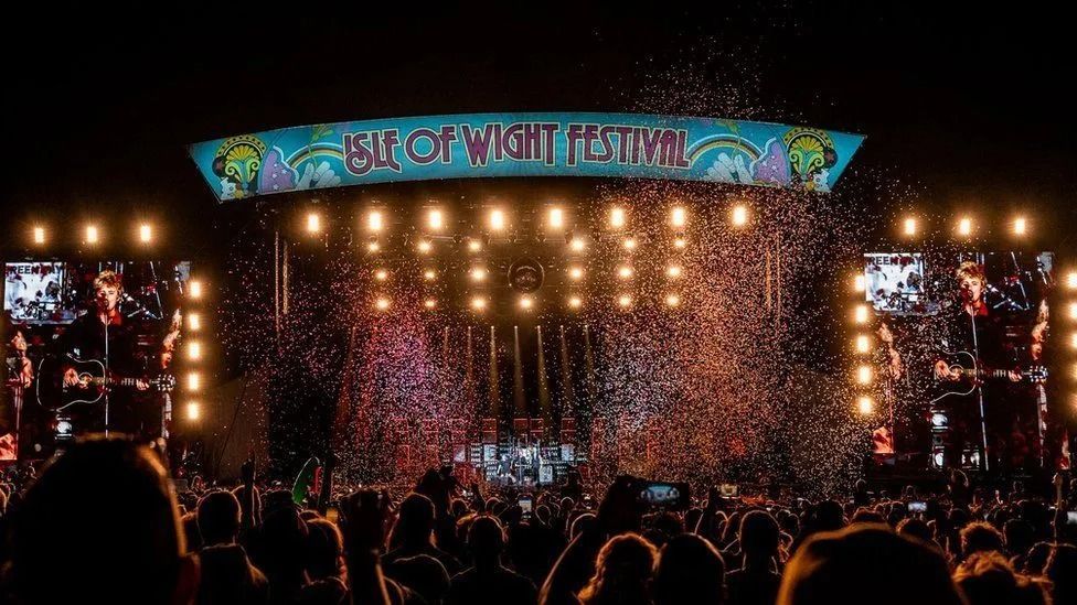 The main stage at the Isle of Wight festival at night time. Confetti is in the air over the heads of crowds of people who are watching the stage. A musician can be seen on the stage holding at guitar.
