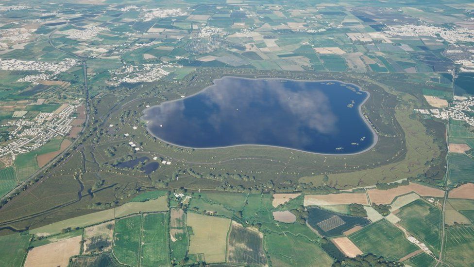 The vast darkblue puddle-shaped reservoir seen from above centred in a patchwork of green and brown greenspace and fields