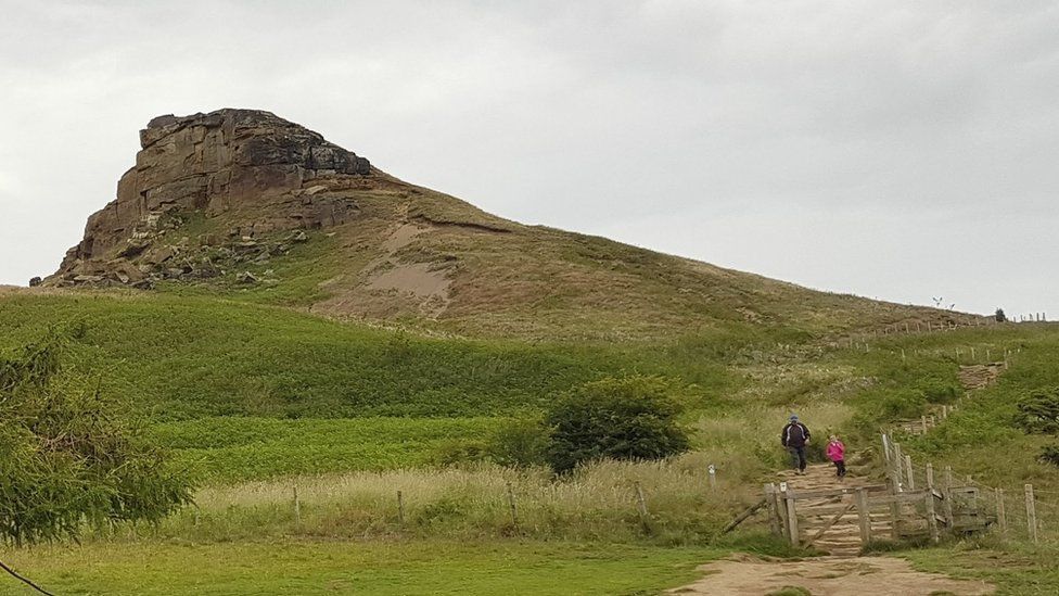 Roseberry Topping is a stone-covered hill surrounded by green fields. People can be seen on a pathway walking toward the viewpoint.