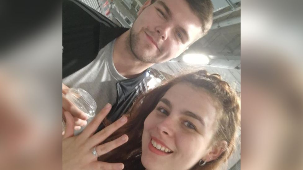 Paul Ward is stood wearing a grey t shirt and adidas jacket, holding a drink, next to Hollie Randall who is holding her hand out in front of her showing a sparkly engagement ring