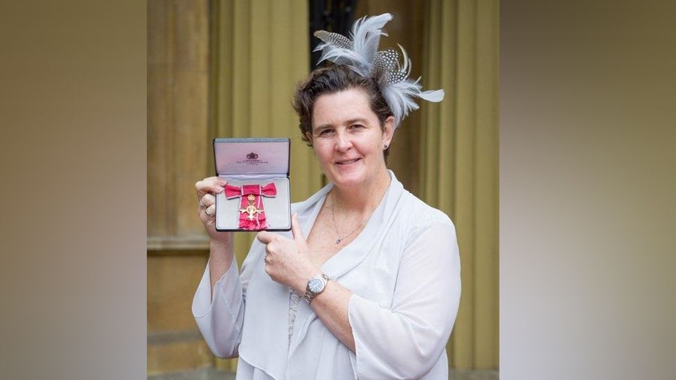 A woman in a white blouse with a feather fascinator holding an award or medal 