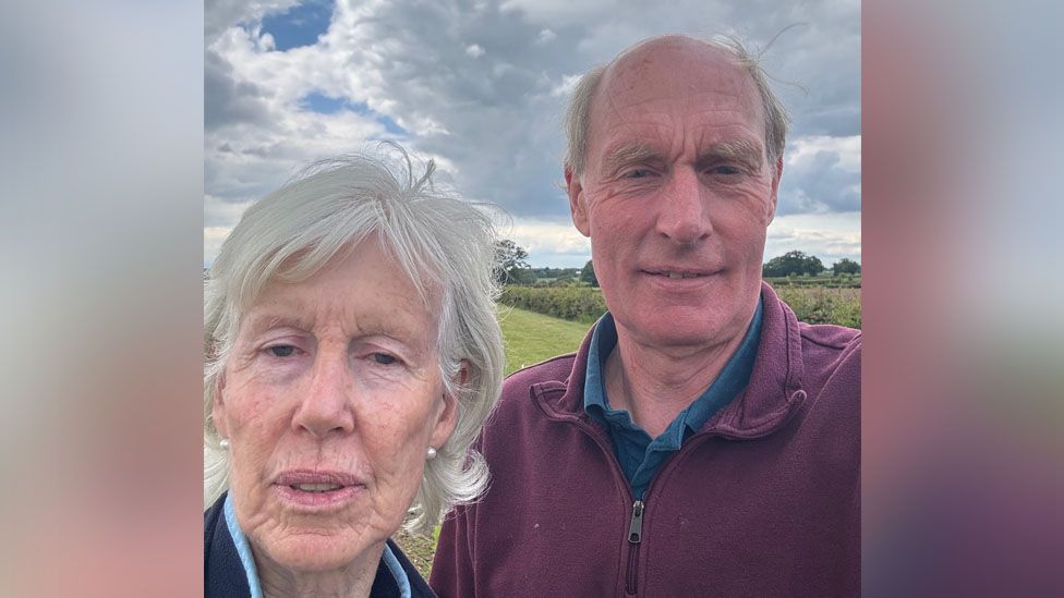 (L-R) A selfie of Jane and Stephen on their farm. Jane has white hair and wearing pearl earrings and Stephen is wearing a quarter zip and a navy shirt underneath. 