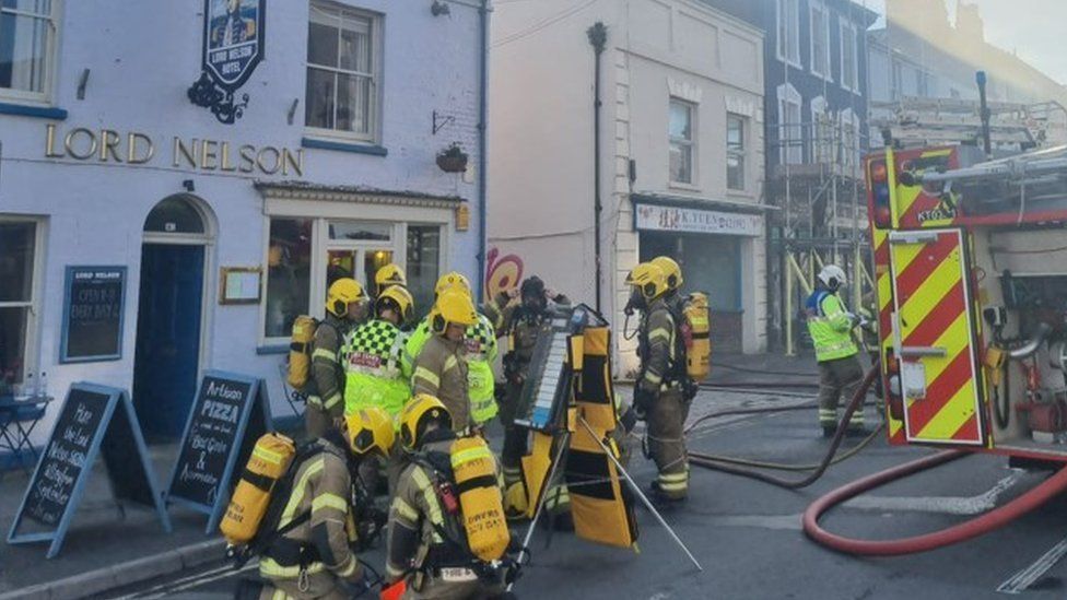 Pub with board signs outside and group of over 10 firefighters some standing others crouching to the side of a smoking building