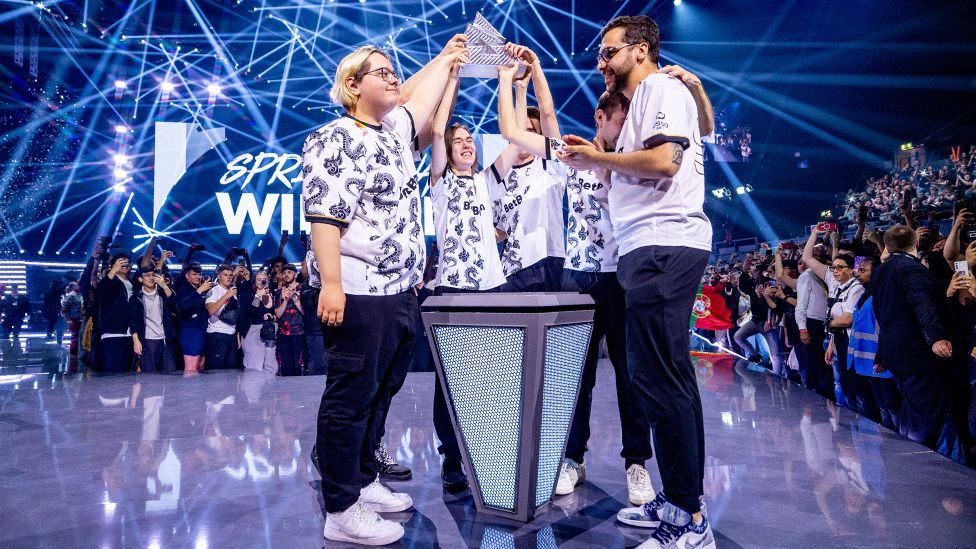 A group of five young people wearing matching white t-shirts with a dragon pattern surround a plinth in the middle of a stage. A cheering crowd looks on as one team member holds a glass, triangular trophy over their head and their team-mates reach out to touch it.