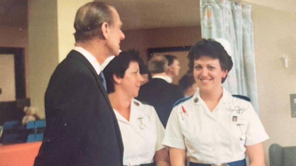 Mary pictured as a young nurse wearing a white nursing uniform which has a blue epaulette on the shoulder and a matching blue belt around her waist. She has short brown hair, a white hat and is smiling at the camera. To her left is Prince Philip wearing a blazer and shirt, he is facing away from the camera but his side profile is visible. 