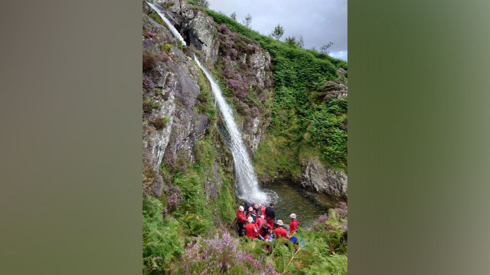 Group of rescuers in red at the bottom of a mountain waterfall, rescuing a man who has fallen from the top